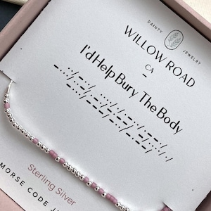 May include: A sterling silver Morse code necklace with a pink and silver bead design. The necklace is displayed on a card that reads "I'd Help Bury The Body" in Morse code, with the Willow Road Jewelry logo.