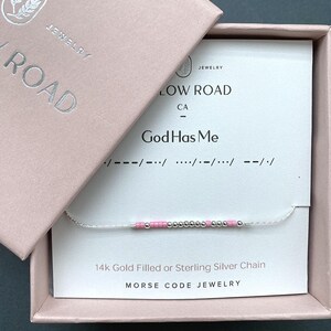 May include: A delicate bracelet featuring a sequence of small pink and silver beads arranged in a Morse code pattern. The bracelet is presented on a white card inside a light pink box, with the text God Has Me and Morse code symbols printed.