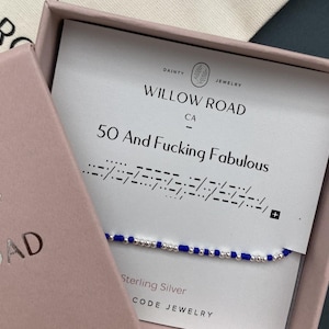 May include: A blue and silver beaded bracelet in a pink gift box. The card inside reads "50 And Fucking Fabulous" with Morse code. The box also displays the brand name "Willow Road CA" and "Sterling Silver Code Jewelry."