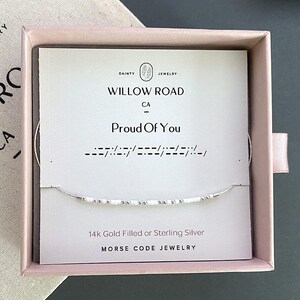 May include: A delicate silver necklace with white beads spelling "Proud Of You" in Morse code, presented in a pink box. The card inside reads "Willow Road CA" and "14k Gold Filled or Sterling Silver Morse Code Jewelry."