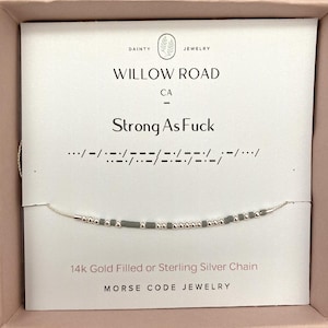 Peut inclure: Un collier délicat en chaîne argentée avec un message en code Morse. La carte indique "Strong As Fuck" avec la traduction en code Morse en dessous. Le collier est présenté dans une boîte rose. Le texte sur la carte dit aussi "14k Gold Filled or Sterling Silver Chain".
