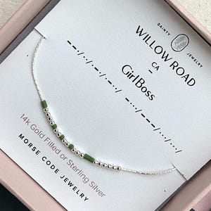 May include: A delicate silver necklace with green and silver beads spelling "Girl Boss" in Morse code. The necklace is displayed on a white card with the words "Willow Road CA" and "Morse Code Jewelry". The jewelry is in a pink box.