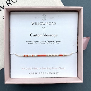 May include: A delicate Morse code bracelet featuring alternating metallic gold-tone beads and vibrant orange beads on a thin chain. The jewelry is displayed on a white card inside a pink box, printed with Willow Road CA and Custom Message.