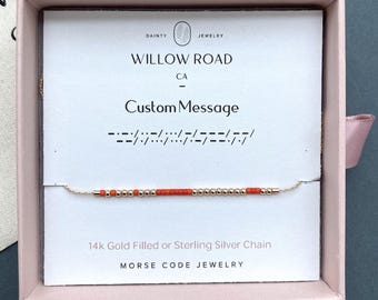 May include: A delicate Morse code bracelet featuring alternating metallic gold-tone beads and vibrant orange beads on a thin chain. The jewelry is displayed on a white card inside a pink box, printed with Willow Road CA and Custom Message.