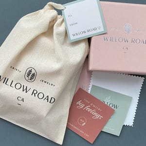 May include: A beige cotton drawstring pouch with the black text Willow Road CA, a light pink jewelry box with silver branding, a light blue gift tag, a white polishing cloth, and two small square cards with the text Tiny Jewelry big feelings.