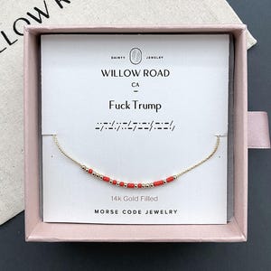 May include: A 14k gold filled Morse code bracelet featuring small red and gold beads on a delicate chain. The jewelry is presented on a white card inside a pink box, with the text "WILLOW ROAD CA", "Fuck Trump", and "MORSE CODE JEWELRY" printed.