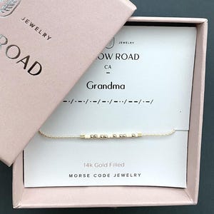 May include: A 14k gold filled Morse code bracelet with alternating cream and gold beads on a thin chain. The bracelet is presented on a white display card labeled "Grandma" with Morse code dots and dashes, inside a light pink jewelry box.