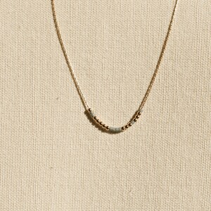 May include: A delicate gold chain necklace featuring a central arrangement of small, round metallic gold beads interspersed with tiny, cylindrical matte grey beads, creating a subtle and refined pattern along the lower curve of the chain.