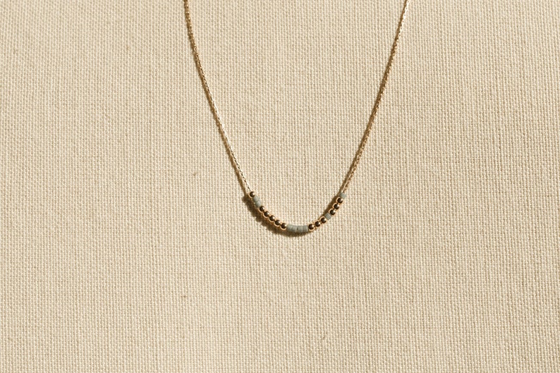 May include: A delicate gold chain necklace featuring a central arrangement of small, round metallic gold beads interspersed with tiny, cylindrical matte grey beads, creating a subtle and refined pattern along the lower curve of the chain.