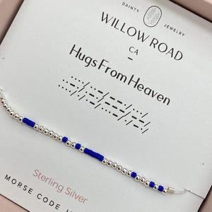 May include: A delicate sterling silver bracelet with blue beads and silver beads. The bracelet is displayed on a card with the text "Hugs From Heaven" and "Willow Road Jewelry CA". The Morse code spells out "Hugs From Heaven".