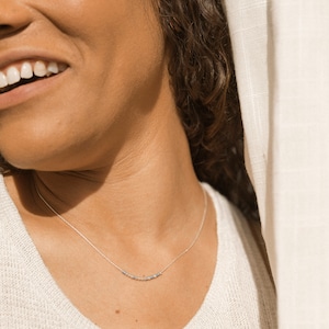 May include: A close-up of a person wearing a white sweater and a silver necklace with small beads. The person is smiling and has curly brown hair.