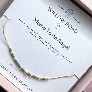 May include: A 14k gold filled bracelet with green and gold beads arranged in Morse code, displayed on a white card labeled Willow Road Jewelry with the text Mama To An Angel. The delicate chain features a repeating pattern of small beads.