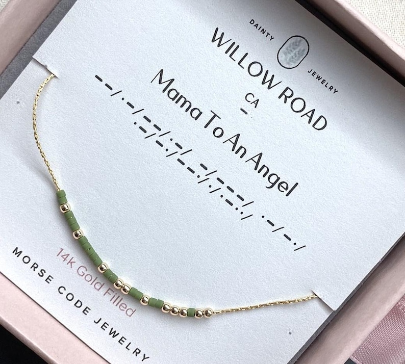 May include: A 14k gold filled bracelet with green and gold beads arranged in Morse code, displayed on a white card labeled Willow Road Jewelry with the text Mama To An Angel. The delicate chain features a repeating pattern of small beads.