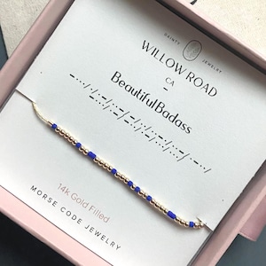 May include: A delicate gold-filled Morse code bracelet with blue beads, presented on a card inside a pink box. The card reads "Beautiful Badass" and "Willow Road Jewelry, CA". The bracelet is 14k gold filled.