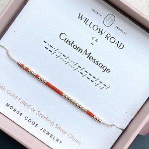 May include: A delicate chain necklace featuring alternating small metallic gold-tone beads and vibrant orange cylindrical beads arranged in a Morse code pattern. The necklace is displayed on a white card labeled Willow Road Jewelry.