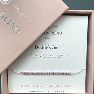 May include: A pink and silver Morse code bracelet in a pink jewelry box. The card reads "Daddy's Girl" with the Morse code translation below. The box also has the text "JEWELRY" and "ROAD". The bracelet is on a silver chain.