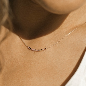 May include: A delicate gold chain necklace featuring a central arrangement of small, polished gold-tone beads interspersed with tiny, pale pink beads. The necklace rests against the skin, showcasing a minimalist design with a thin, refined metal link chain.