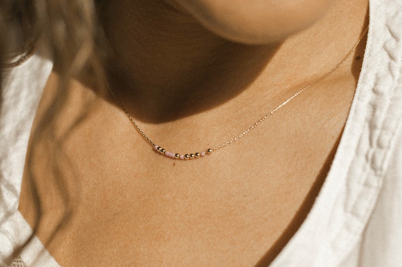May include: A delicate gold chain necklace featuring a central arrangement of small, polished gold-tone beads interspersed with tiny, pale pink beads. The necklace rests against the skin, showcasing a minimalist design with a thin, refined metal link chain.