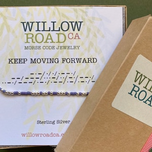 May include: A small box with a light brown lid and a pink ribbon. The box has a white label with the text "WILLOW ROAD CA" and "MORSE CODE JEWELRY". The box is sitting on a green surface.