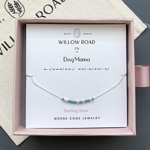 May include: A sterling silver chain bracelet featuring a series of small blue and silver beads arranged in a Morse code pattern. The bracelet is displayed on a white card labeled Willow Road CA Dog Mama inside a light pink gift box.