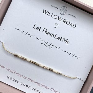 May include: A delicate gold-toned chain bracelet featuring a sequence of small metallic gold and cream-colored beads arranged in a Morse code pattern. The bracelet is displayed on a white card printed with the text Willow Road, Let Them Let Me.