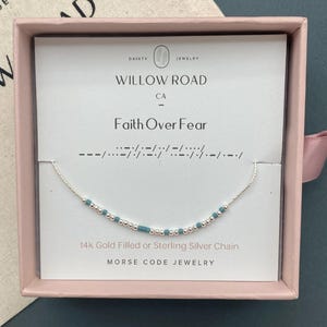 May include: A silver necklace with turquoise beads spelling out "Faith Over Fear" in Morse code. The necklace is displayed in a pink box with the words "Willow Road" and "Morse Code Jewelry" printed on the card.
