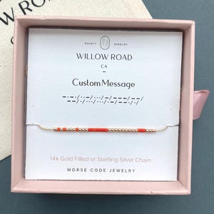 May include: A gold-coloured necklace with red and gold beads, presented in a pink box. The necklace is part of the Morse Code Jewelry collection. The card inside the box reads "WILLOW ROAD CA Custom Message" and "14k Gold Filled or Sterling Silver Chain".