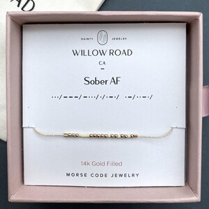 May include: A 14k gold filled Morse code bracelet featuring small metallic gold and cream-colored beads on a thin cord. The bracelet is displayed on a white card inside a pink box, with the text "WILLOW ROAD CA" and "Sober AF" printed above.
