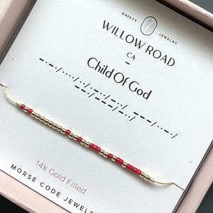 May include: A 14k gold filled Morse code bracelet featuring alternating small red and metallic gold beads on a thin cord. The bracelet is displayed on a white card printed with the text Willow Road, Child Of God, and 14k Gold Filled.