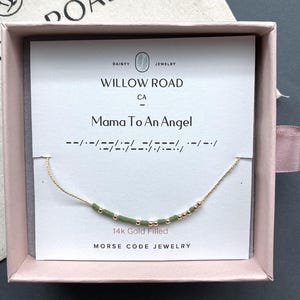 May include: A 14k gold filled Morse code necklace featuring alternating small gold beads and sage green cylindrical beads on a delicate gold chain, presented on a white display card labeled Willow Road with the text Mama To An Angel.