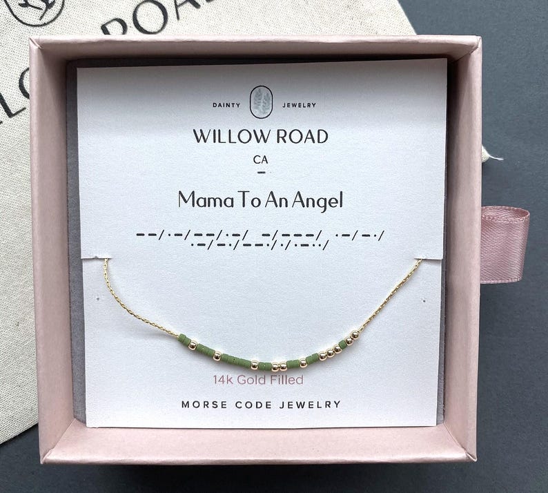 May include: A 14k gold filled Morse code necklace featuring alternating small gold beads and sage green cylindrical beads on a delicate gold chain, presented on a white display card labeled Willow Road with the text Mama To An Angel.