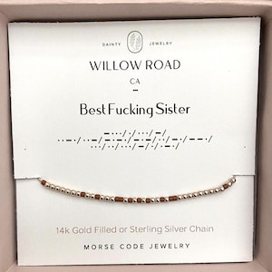 May include: A gold-colored beaded bracelet with brown beads spelling out a message in Morse code. The card reads "Best Fucking Sister" with "Willow Road CA" above. The text "14k Gold Filled or Sterling Silver Chain" is also visible.