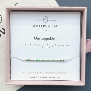 May include: A delicate chain bracelet featuring alternating small silver-toned beads and light green cylindrical beads arranged in a Morse code pattern. The bracelet is displayed on a white card inside a pink box, with the text Unstoppable printed above.