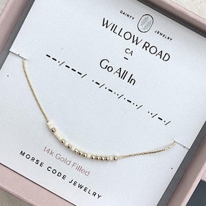 May include: A 14k gold filled Morse code necklace featuring a delicate gold chain with alternating small white and gold beads. The necklace is displayed on a white card printed with the text Willow Road, Go All In, and Morse code symbols.