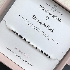 Peut inclure: Un bracelet délicat composé de perles argentées et noires alternées formant un code Morse. Le bracelet est présenté sur une carte blanche portant l'inscription Willow Road Jewelry et Strong As Fuck, dans une boîte rose pâle.