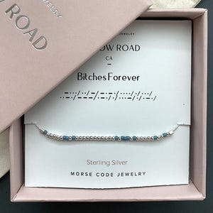 May include: A sterling silver Morse code bracelet featuring small metallic silver beads and muted blue-grey beads on a thin cord. The bracelet is displayed on a white card inside a light pink box, with the text Bitches Forever printed above.