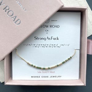 May include: A 14k gold filled Morse code necklace with alternating gold beads and olive green cylindrical beads on a thin gold chain. The necklace is displayed on a white card inside a pink box, featuring the text Strong As Fuck and Morse code.