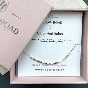 May include: A gold-filled or sterling silver Morse code necklace with white and gold beads, presented in a pink box. The card inside reads "Clean And Sober" with the Morse code equivalent. The box also displays "JEWELRY" and "ALLOW ROAD CA".
