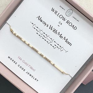 May include: A 14k gold filled Morse code bracelet featuring alternating small gold beads and cream-colored cylindrical beads. The bracelet is displayed on a white card printed with the text Willow Road Dainty Jewelry and Always With Me Mom.