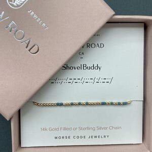 May include: A gold-colored beaded bracelet with teal beads, presented in a pink jewelry box. The box is labeled "Jewelry Road" and "Shovel Buddy". The bracelet is described as 14k Gold Filled or Sterling Silver Chain, Morse Code Jewelry.