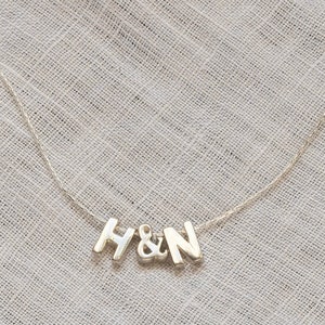 May include: A silver necklace with the letters "H&N" in a block font. The letters are connected by a delicate chain, set against a textured, light gray background.