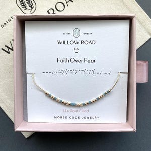 May include: A delicate gold-filled chain bracelet featuring small metallic gold and light blue beads arranged in a Morse code pattern. The bracelet is displayed on a white card printed with the text Willow Road, Faith Over Fear, and 14k Gold Filled.