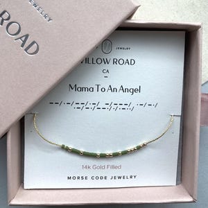May include: A 14k gold filled Morse code bracelet with green and gold beads on a white display card reading Mama To An Angel, housed in a light pink gift box labeled Willow Road Jewelry. The delicate chain features a sequence of beads.