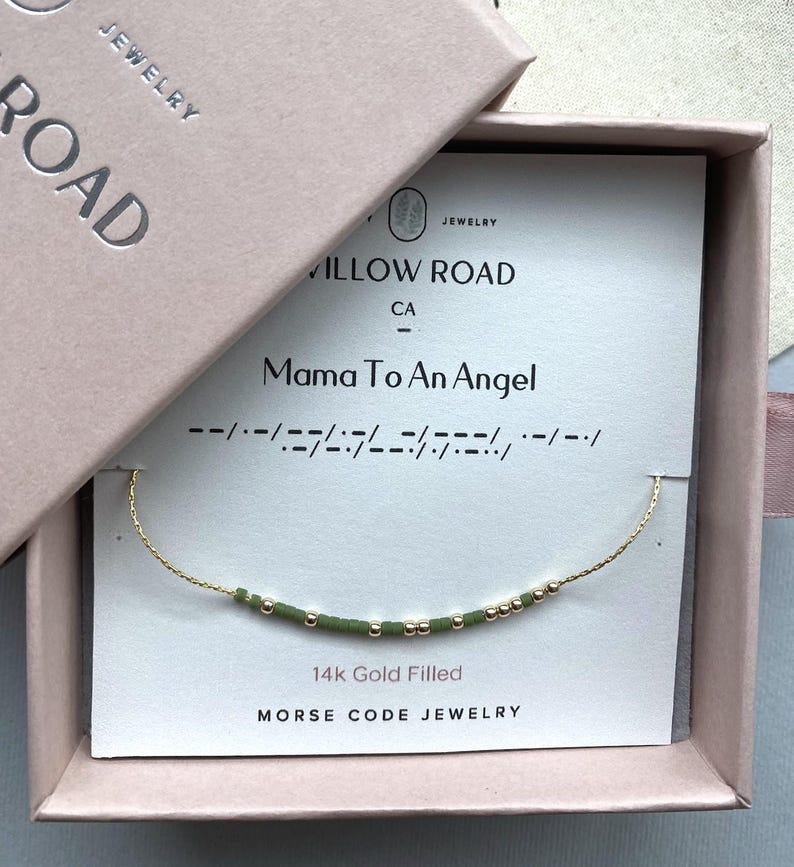 May include: A 14k gold filled Morse code bracelet with green and gold beads on a white display card reading Mama To An Angel, housed in a light pink gift box labeled Willow Road Jewelry. The delicate chain features a sequence of beads.