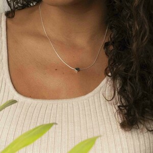 May include: A delicate silver necklace with a small, heart-shaped pendant. The pendant is a dark color, possibly black or gray, and hangs from a thin chain. The necklace is worn against a light-colored top.