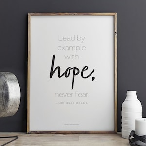 Peut inclure: Impression encadrée avec la citation "Lead by example with hope, never fear" de Michelle Obama. Le texte noir est sur fond blanc, dans un cadre en bois. Des objets décoratifs sont visibles sur une surface en bois.