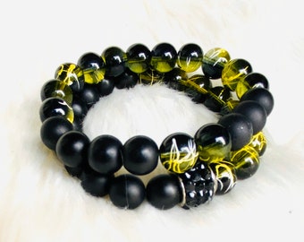 May include: Two bracelets, one black and one with green and yellow beads. The green and yellow beads have a white swirl pattern. The black bracelet has a black rhinestone charm.