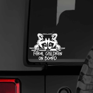 May include: White vinyl decal of a raccoon peeking over a horizontal banner with the text 'Feral Children On Board'. The decal is affixed to the rear of a black Jeep. The Jeep's tire is partially visible in the lower right corner of the image. The decal is a humorous design suitable for parents of mischievous children.