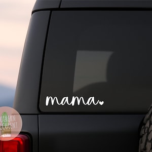 May include: A white vinyl decal applied to a dark vehicle window. The decal features the word mama in a cursive script font, followed by a small heart symbol. The design is clean, simple, and adheres directly to the glass surface of the car.