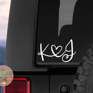 May include: White vinyl decal with the letters "K & J" with a heart symbol between the letters. The decal is on the back window of a black SUV.